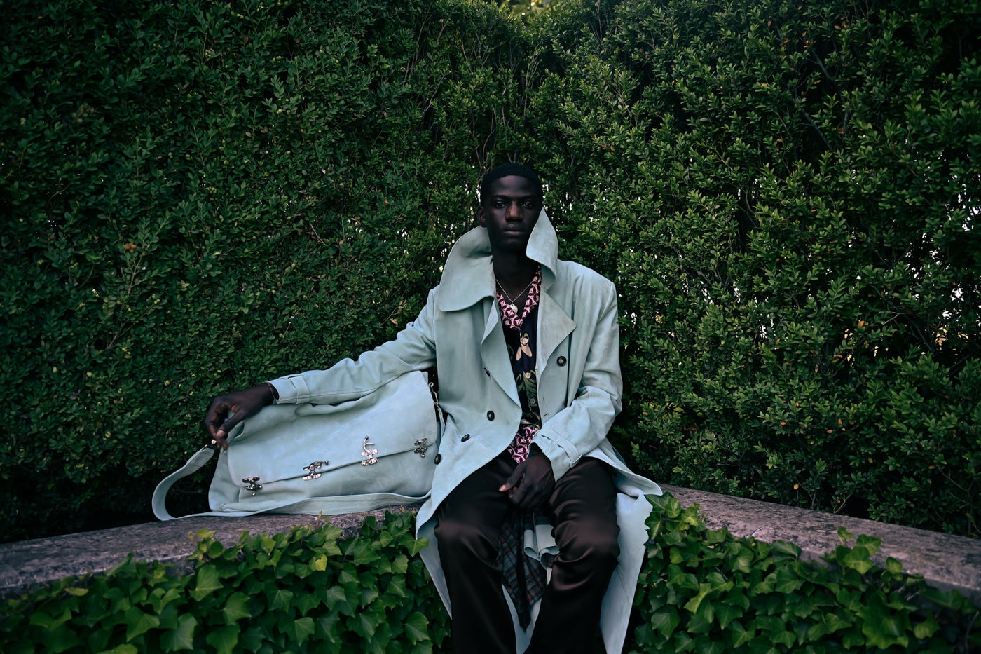 Pierre Blanc Autumn Winter 2026-27 campaign - pale trench coat with a leather holdall, seated against garden hedging